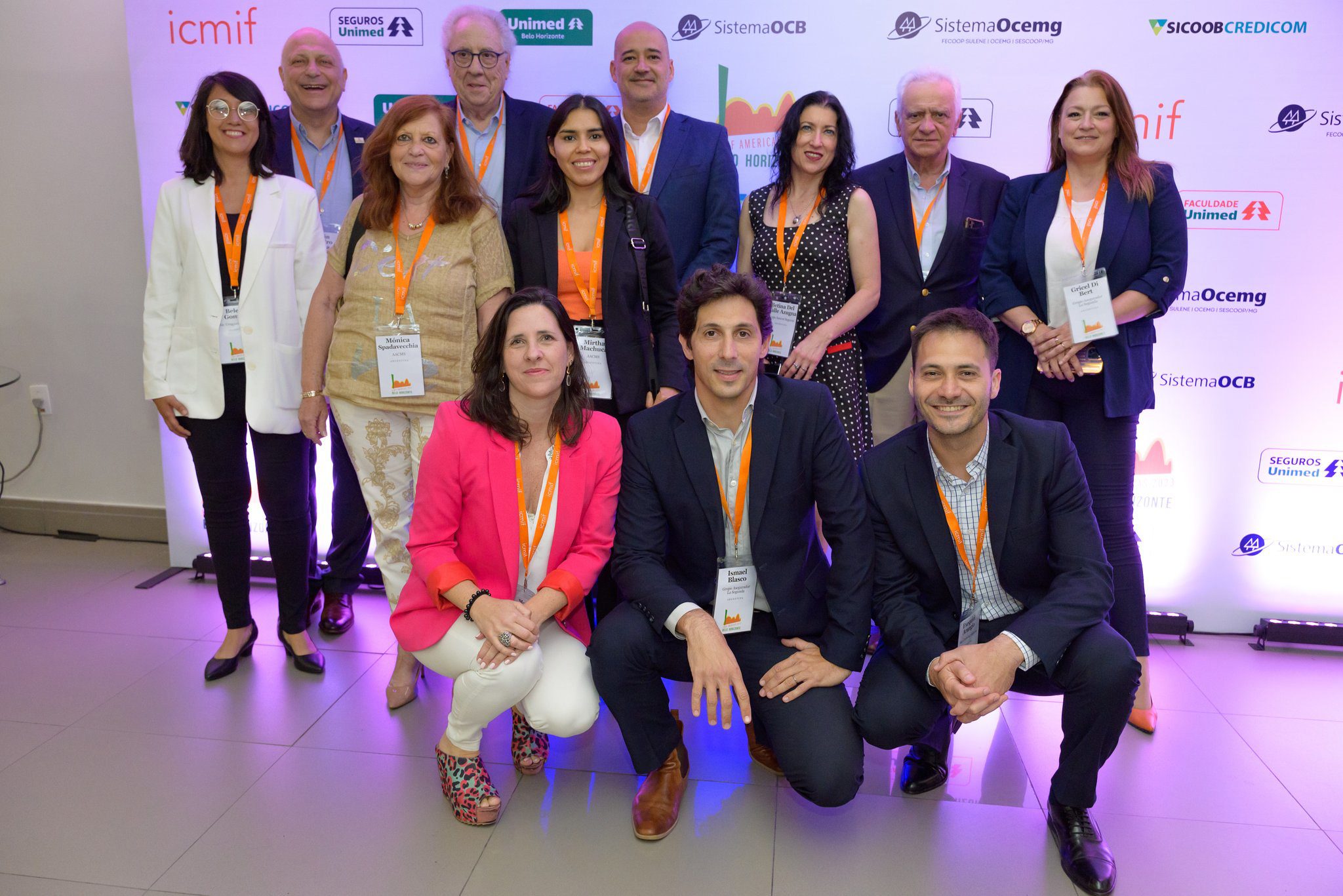 A group of thirteen people, dressed in business attire with conference badges and orange lanyards, pose and smile together in front of a white backdrop with various sponsor logos.