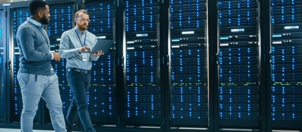 Two men walk through a server room with rows of tall, black server racks filled with blue and green lights. One holds a laptop and they appear to be discussing or checking the servers.