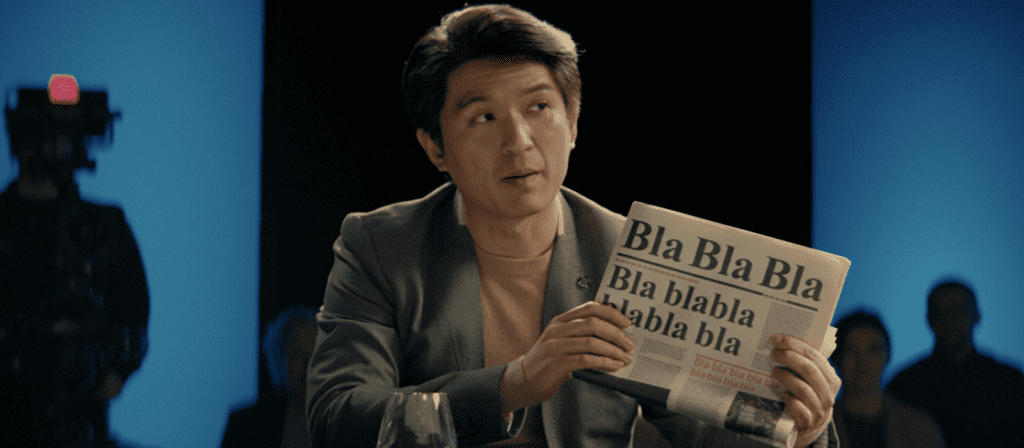 A man in a suit holds up a newspaper that reads “Bla Bla Bla” in large text, sitting at a table in a studio with people and a camera in the background.