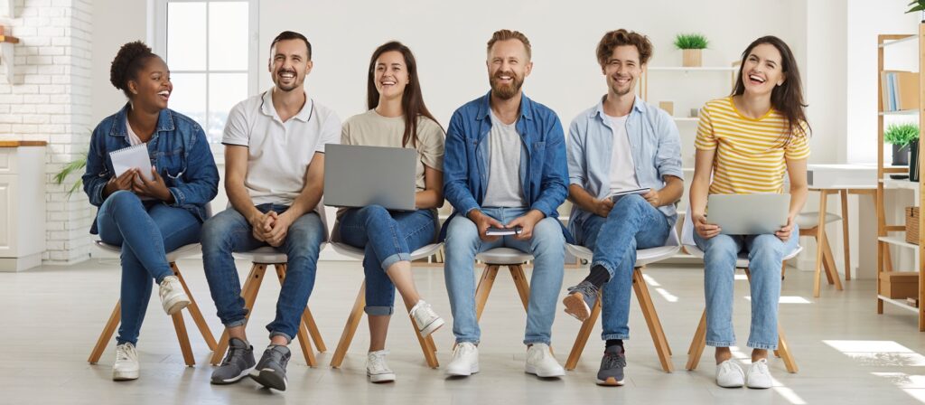 Six young adults sit in a row on chairs, smiling and holding laptops, notebooks, or tablets in a bright, modern room with plants and shelves in the background.