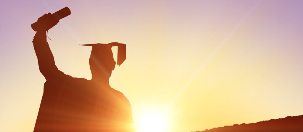 A silhouette of a graduate wearing a cap and gown raises a diploma in the air, standing in front of a bright sunrise or sunset.