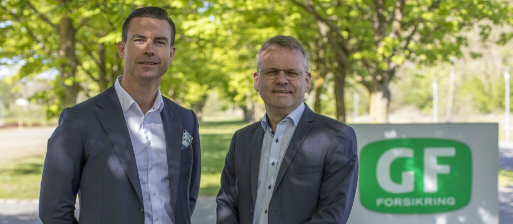 Two men in suits stand outdoors in front of a green GF Forsikring sign, with leafy trees and sunlight in the background.