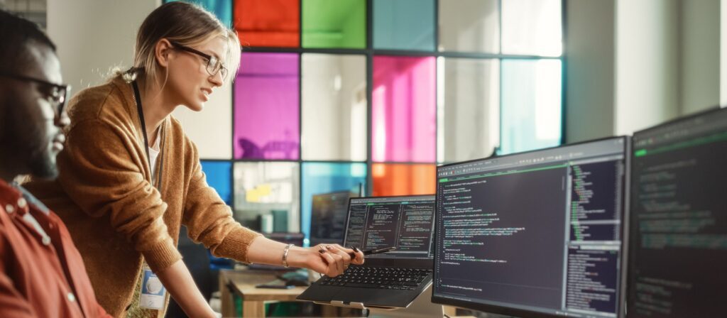 Two people collaborate in a modern office. One person stands pointing at code on a laptop screen, while the other sits watching. Large monitors and colorful glass panels are visible in the background.