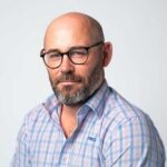 Bald man with a beard and glasses, wearing a light blue and pink plaid shirt, looking at the camera with a neutral expression against a plain light background.