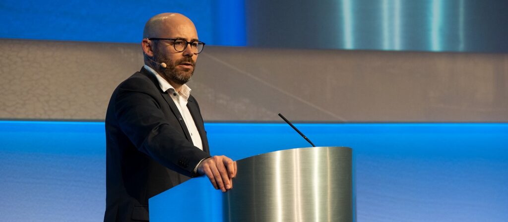 A man in a suit and glasses speaks into a headset microphone while standing at a podium on a stage with blue lighting.