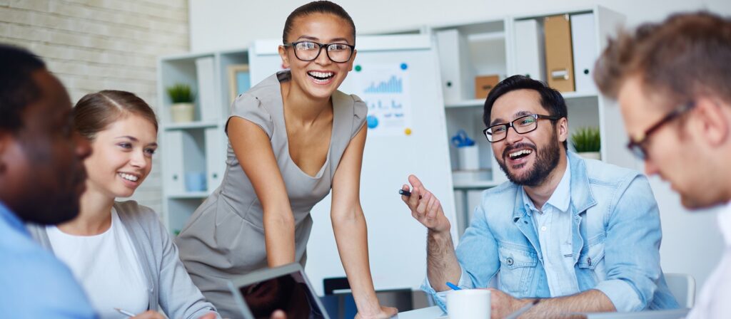Five people in a modern office are gathered around a table, smiling and laughing while engaged in a group discussion. One woman stands, leaning forward, while the others are seated, appearing enthusiastic and collaborative.