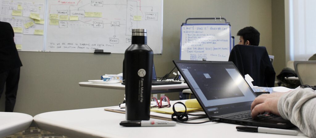 A person types on a laptop at a table with a water bottle, glasses, and marker; in the background, people work near large whiteboards covered with notes and diagrams.