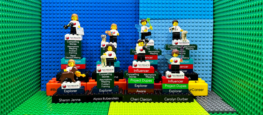 A display of six LEGO minifigures standing on colorful brick podiums labeled with names and words like Explorer, Project Dupe, and Influencer, against a blue and green LEGO backdrop.