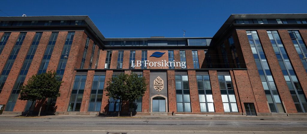 A large brick building with tall glass windows and the sign LB Forsikring on the facade, set against a clear blue sky. Two small trees line the street in front of the building.
