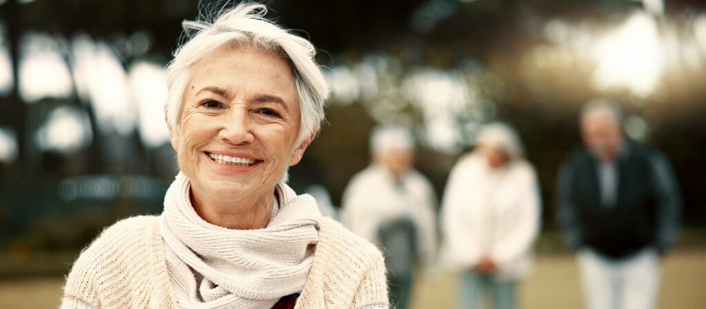 Smiling older woman wearing a scarf and sweater stands outdoors, with three other older adults blurred in the background, suggesting a group gathering in a park or garden.