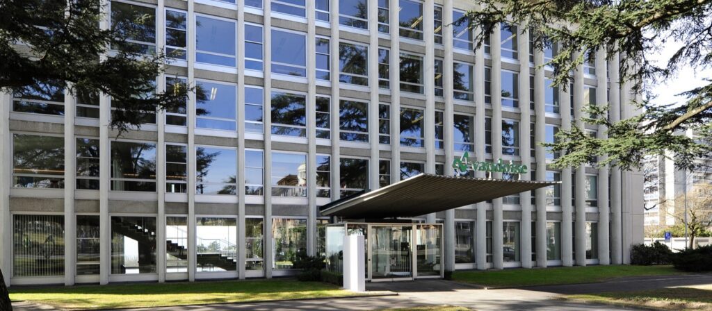 A modern office building with large glass windows and a covered entrance. The word Randstad is displayed above the entrance. Trees and green grass are visible in the foreground.