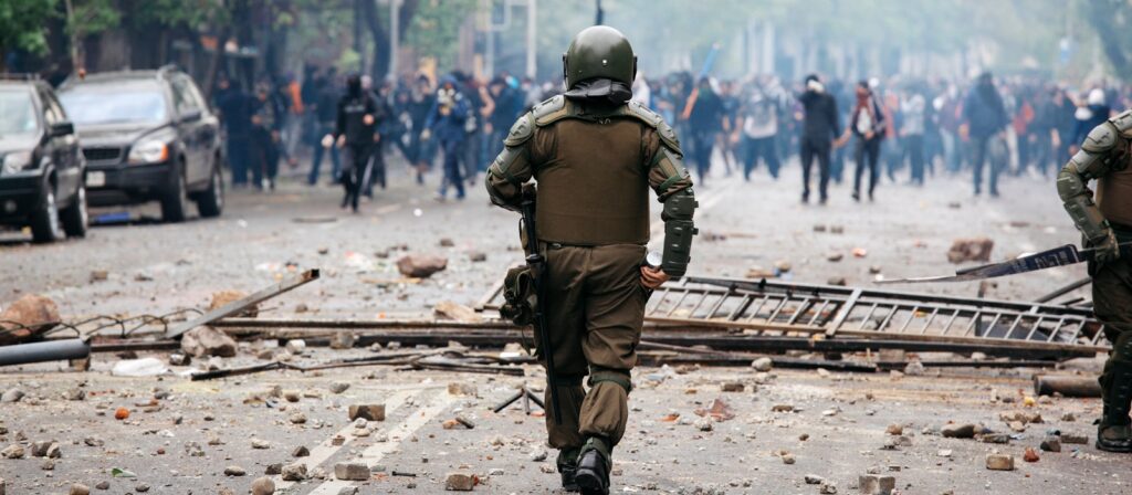 A police officer in riot gear walks toward a crowd of protesters on a debris-strewn street, surrounded by smoke and scattered objects, with damaged vehicles visible in the background.