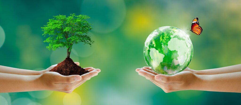Two hands hold soil with a small tree on the left, and another hand holds a glass globe with a butterfly perched on top, symbolizing environmental protection and sustainability on a green, blurred background.