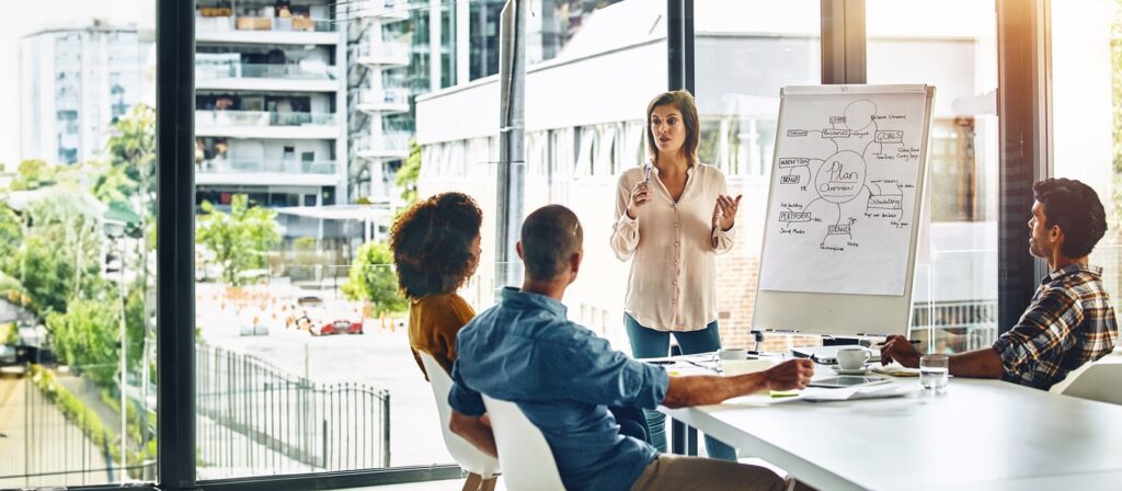A woman stands presenting near a flip chart with a mind map, while three colleagues sit at a table in a modern office with large windows and an urban view outside.