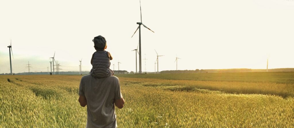 A man carrying a child on his shoulders stands in a field of tall grass, facing several wind turbines on a bright, sunny day. The sky is clear and the landscape is open and peaceful.