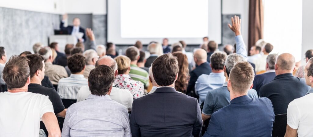 A group of people sit facing a screen and speaker in a conference room. Several audience members have raised their hands, suggesting participation or questions during a presentation.