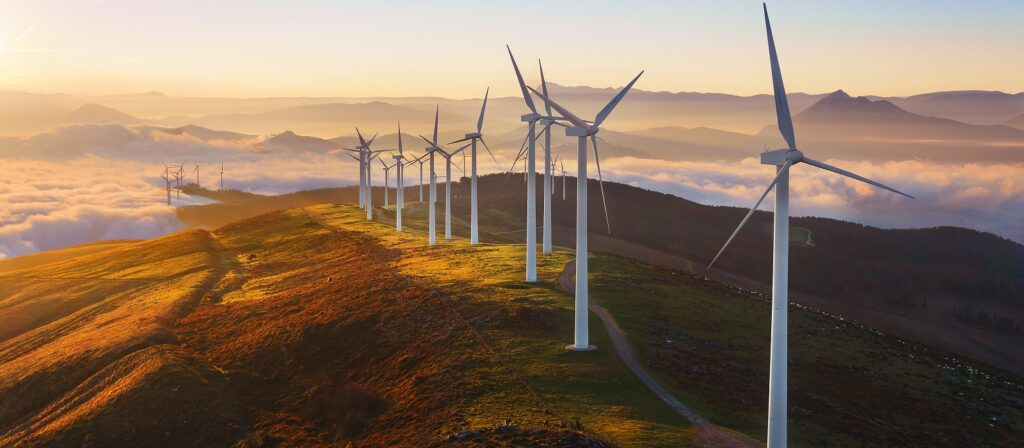 A row of wind turbines stands on a grassy hill at sunrise, with clouds below and mountains in the background. The sunlight casts a warm glow over the landscape.