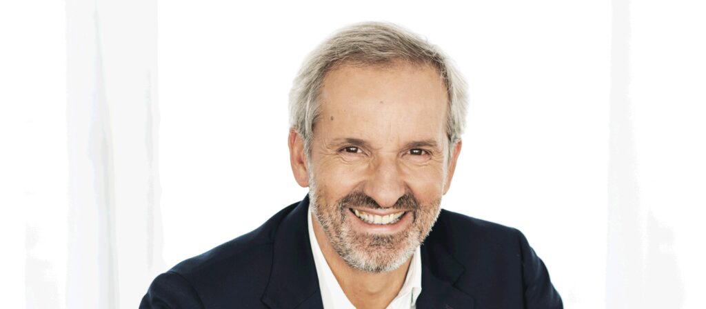 A middle-aged man with gray hair and a beard is smiling warmly. He is wearing a dark blazer over a white shirt and is posed against a bright, white background.