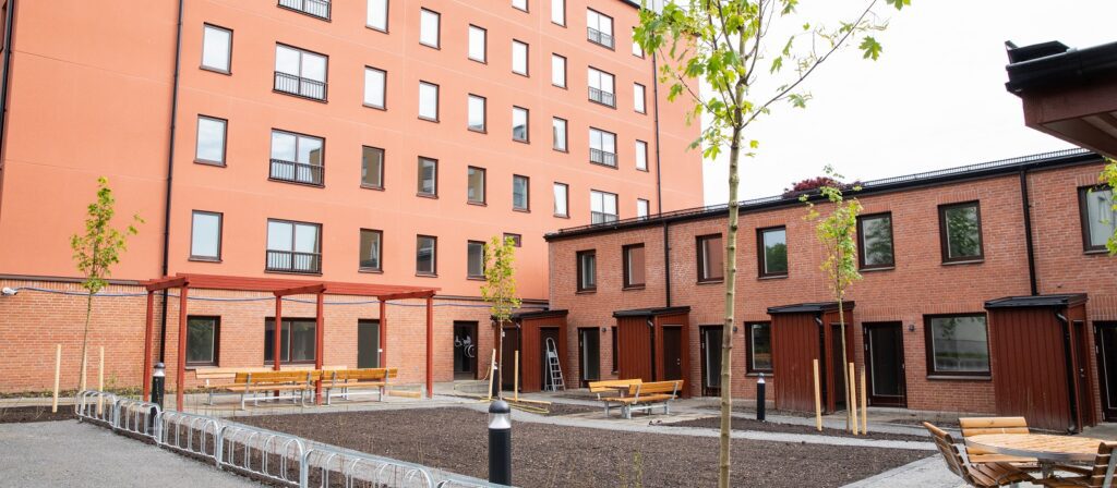 A modern courtyard with young trees, several wooden benches, bike racks, and two adjoining brick buildings, one three stories and the other six stories high, with many windows.