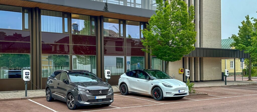 Two electric cars, one gray and one white, are parked side by side at charging stations in front of a modern glass building with trees and greenery nearby.