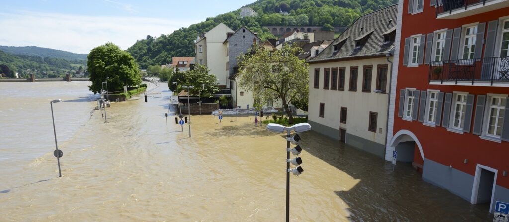 Floodwaters submerge streets and buildings in a town near a river, with muddy water reaching halfway up doorways. Trees and lampposts are partially underwater, and green hills are visible in the background.
