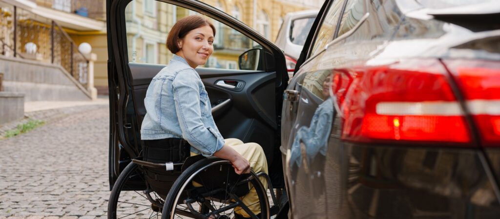 A woman in a wheelchair smiles while getting into a car parked on a cobblestone street, with buildings in the background.
