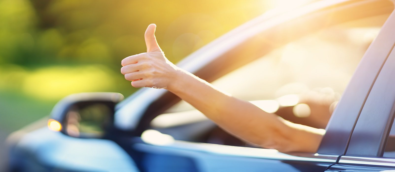 A person’s hand sticks out of a car window giving a thumbs-up gesture, with sunlight and greenery blurred in the background.
