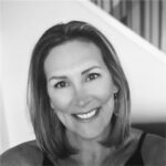 A woman with shoulder-length hair smiles at the camera. She is wearing earrings and a sleeveless top. The background features a staircase and is softly out of focus. The image is in black and white.