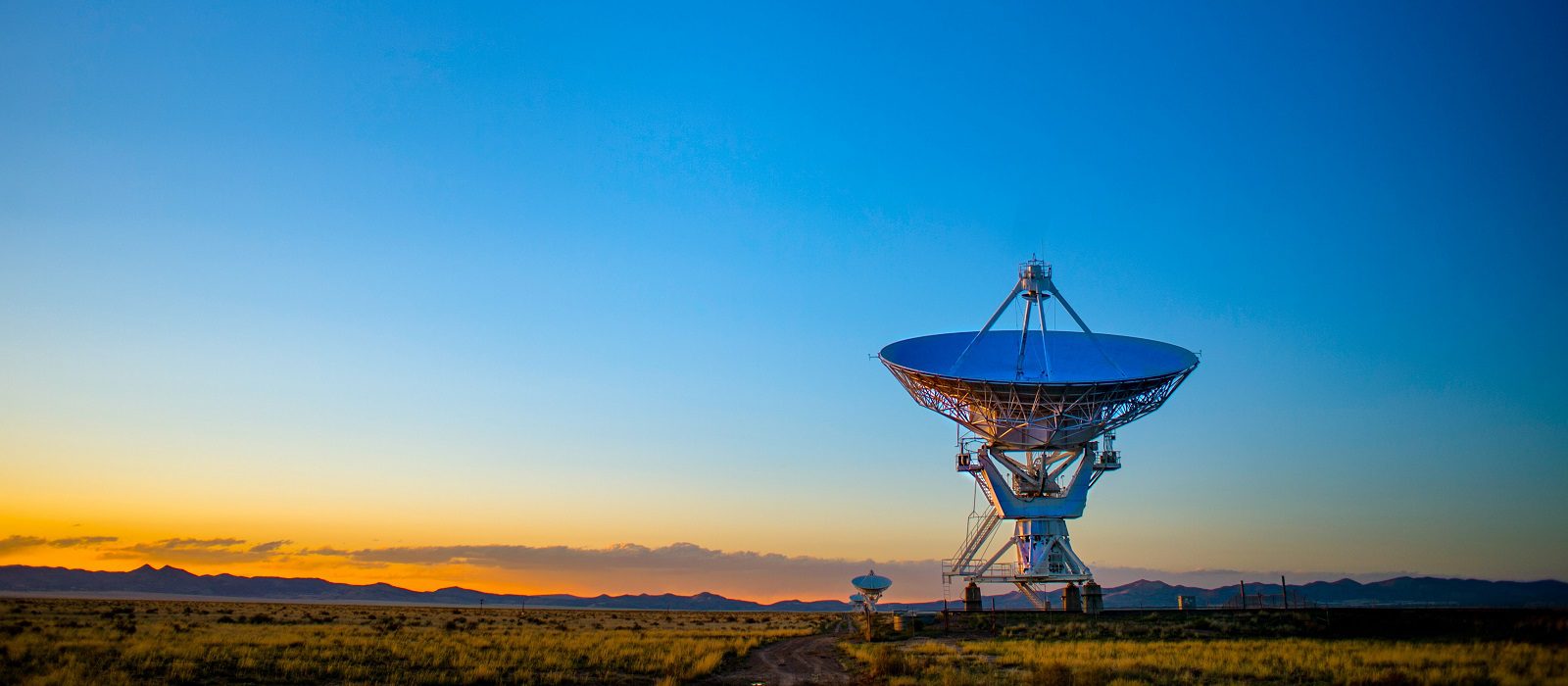 A large radio telescope dish stands in an open field at sunset, with a clear blue and orange sky and distant mountains on the horizon.
