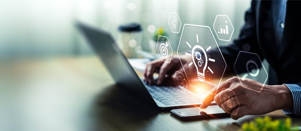 A person in business attire uses a laptop and smartphone at a desk, with digital icons like a lightbulb, gears, and graphs overlaid, symbolizing innovation and technology.
