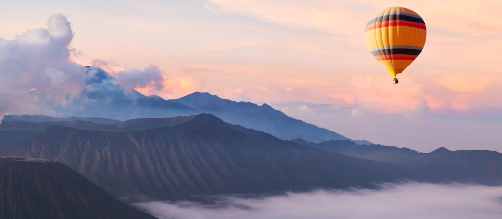 A colorful hot air balloon floats above misty mountains at sunrise, with soft clouds and pastel hues in the sky.