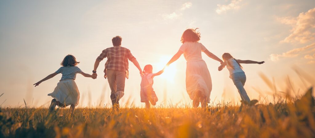 A family of five, holding hands, runs through a grassy field toward the sunset, with the sky glowing warmly and their silhouettes highlighted by the sunlight.