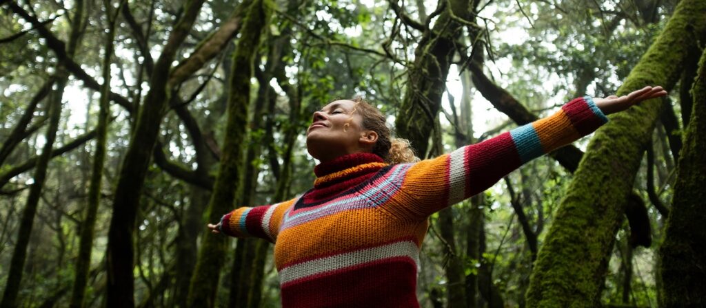 A person in a colorful striped sweater stands in a lush, green forest with arms outstretched and head tilted up, appearing joyful and at peace among tall mossy trees.