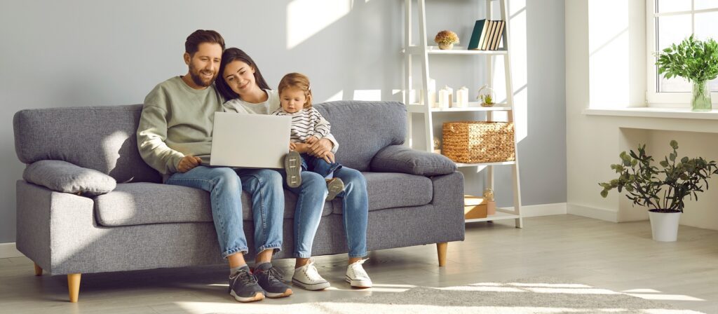 A man, woman, and young child sit together on a gray sofa, smiling and looking at a laptop. Sunlight brightens the modern living room with plants and shelves in the background.