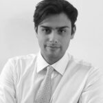 A man with dark hair wearing a white shirt and patterned tie looks at the camera with a neutral expression against a plain background. The image is in black and white.