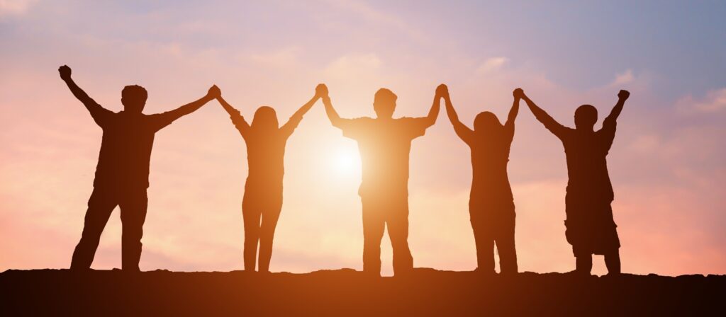 Five people stand in a row holding hands with arms raised, silhouetted against a colorful sunset sky, symbolizing unity and teamwork.
