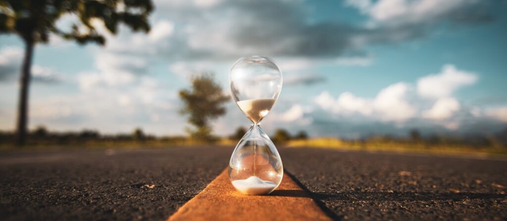 A glass hourglass with sand flowing inside sits centered on an empty road, with blurred trees and sky in the background on a sunny day.