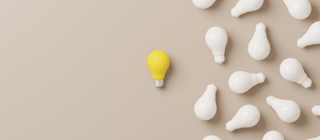 A group of white light bulbs lays scattered on a beige surface, with a single yellow light bulb standing out in the center left, separated from the cluster on the right.
