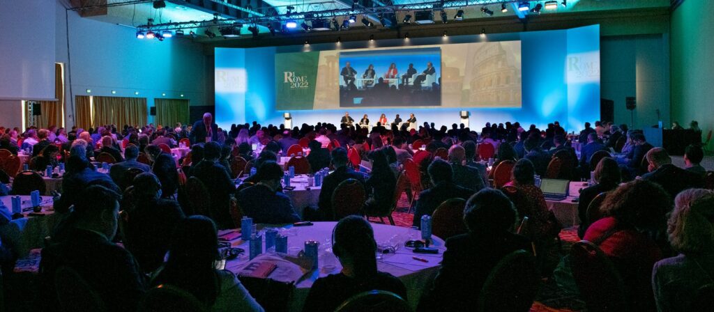 A large audience sits at round tables in a dimly lit conference hall, watching a panel discussion on a brightly lit stage with multiple speakers and a projected event backdrop.