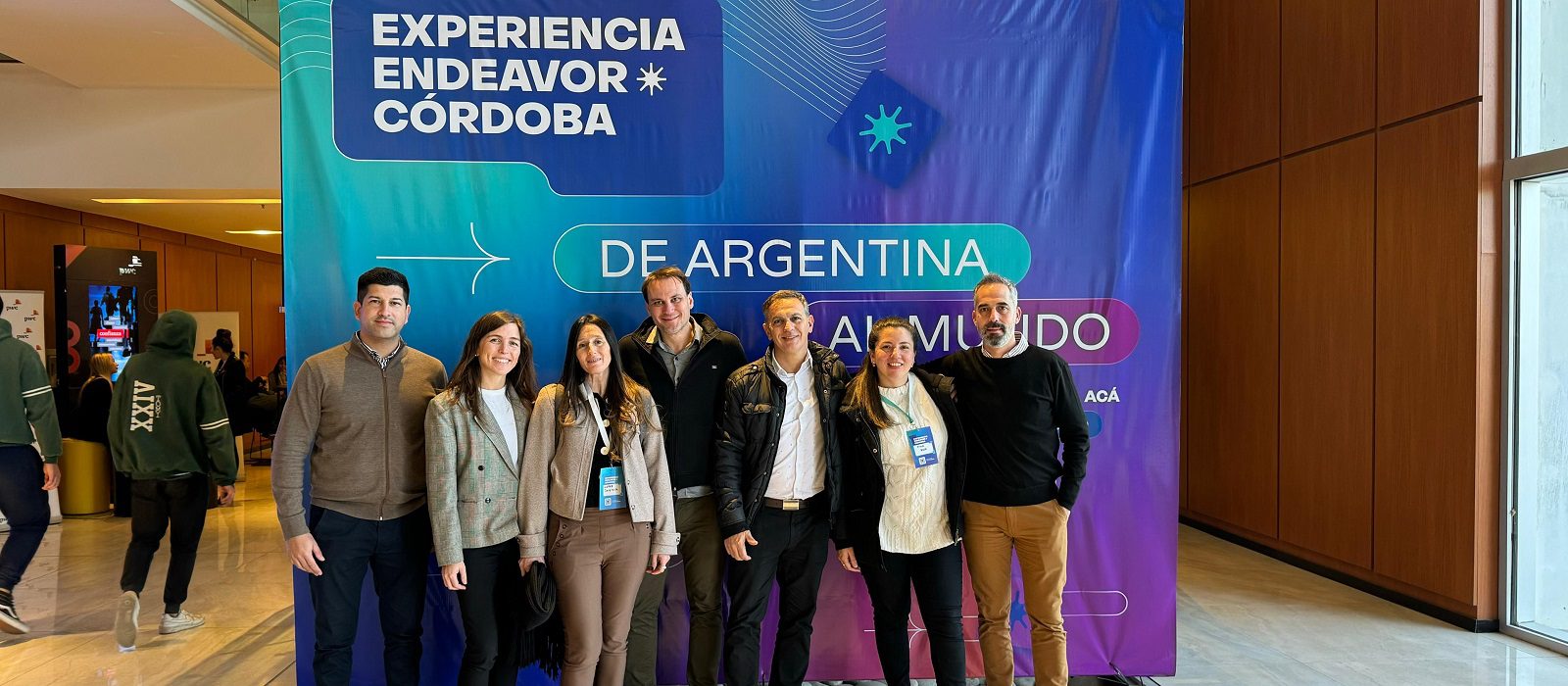 Seven people stand smiling in front of a colorful event banner that reads Experiencia Endeavor Córdoba, De Argentina al Mundo in a modern indoor setting with wooden walls.