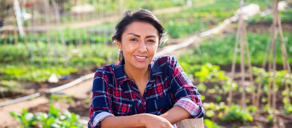 A woman wearing a red and blue plaid shirt smiles while leaning on a fence in a lush vegetable garden with green plants and wooden stakes in the background.