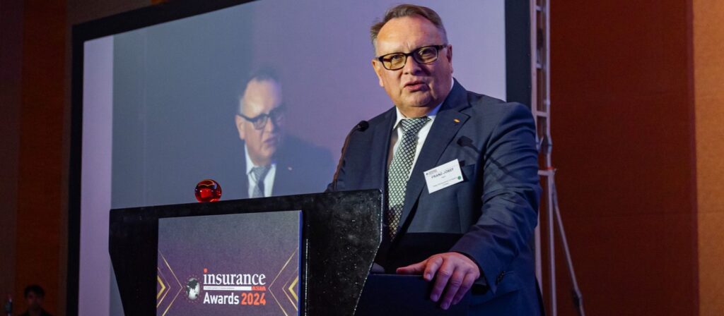 A man in a suit and glasses speaks at a podium marked Insurance Awards 2024, with his image projected on a screen behind him at an indoor event.