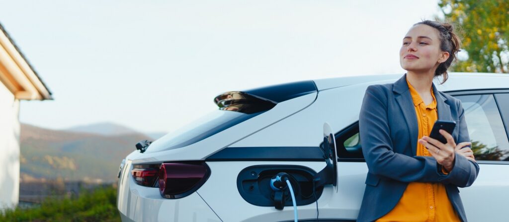 A woman in business attire stands confidently next to a white electric car that is charging. She holds a smartphone and smiles, with mountains and a house visible in the background.
