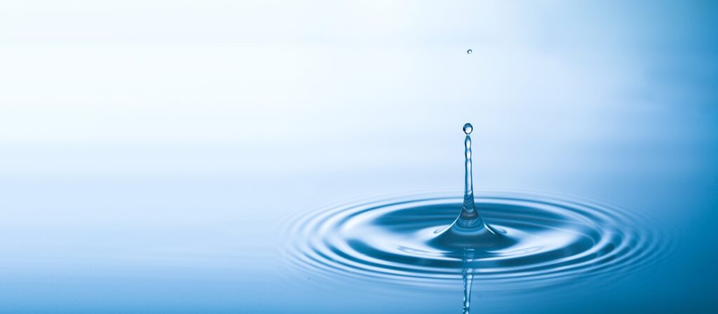 A water droplet splashes into a calm surface, creating ripples in the blue water with a blurred white background.