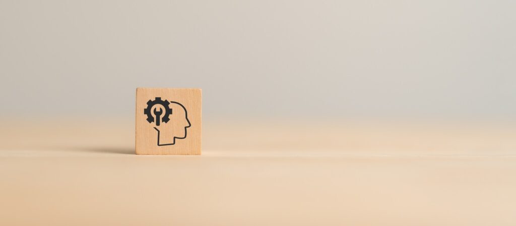 A wooden block with a silhouette of a head and a gear inside it, symbolizing thinking or problem-solving, placed on a plain light surface with a neutral background.