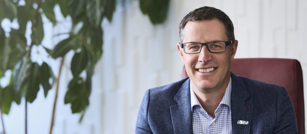 A smiling man with short brown hair and glasses, wearing a blue blazer and checkered shirt, sits in a red chair near a leafy plant and a white textured wall.