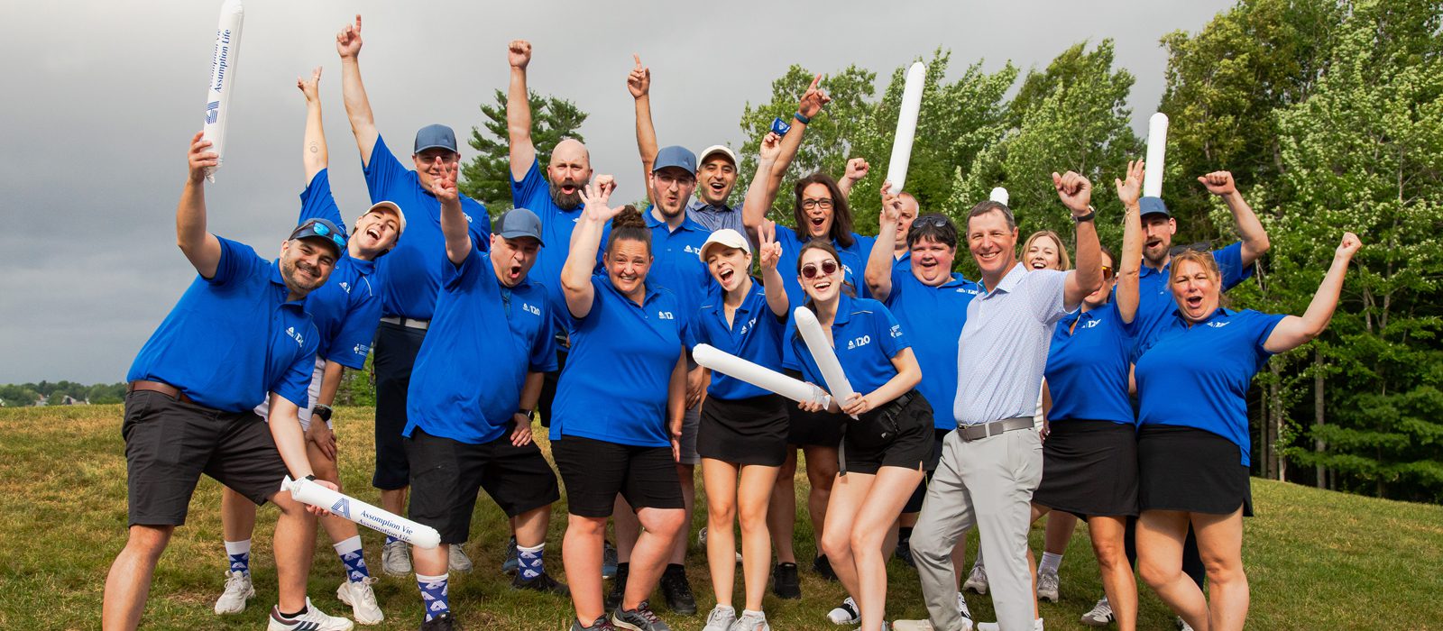 A group of people in matching blue shirts and hats cheer and celebrate outdoors, some holding inflatable sticks, with trees and cloudy sky in the background.