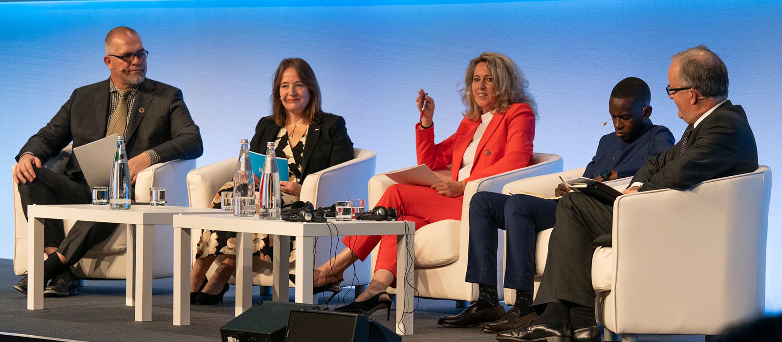 Five people in business attire sit on a stage panel, having a discussion. Three men and two women are seated on white chairs around a white table with microphones, water bottles, and papers, against a blue-lit background.