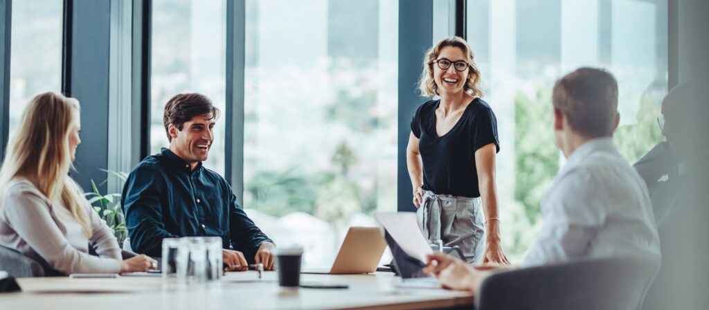 Four colleagues sit around a conference table in a bright office. A woman stands smiling and talking, while the others listen and laugh, suggesting a positive, collaborative meeting.