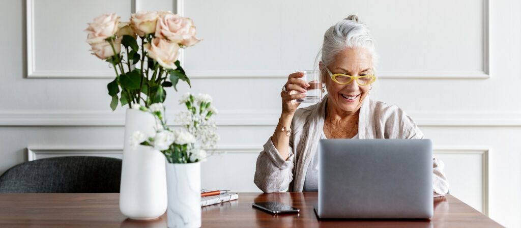 Smiling older woman with gray hair and yellow glasses sits at a table with a laptop, holding a glass of water. Vases with flowers and a smartphone are also on the table in front of her.